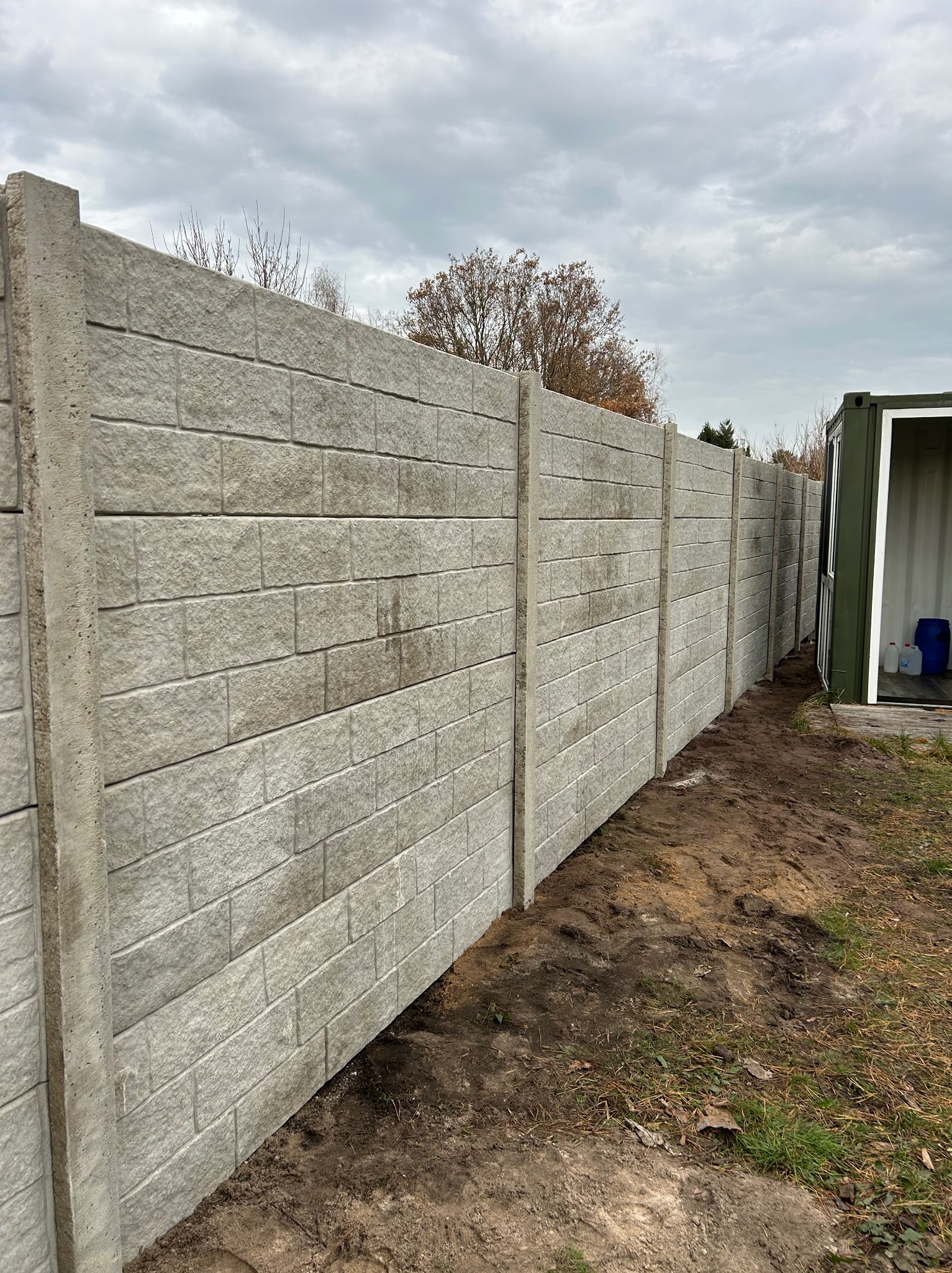 Długi, szary mur z prefabrykatów betonowych, imitujących cegłę, z widocznymi słupkami, stojący na granicy działki, z kontenerem magazynowym w tle.