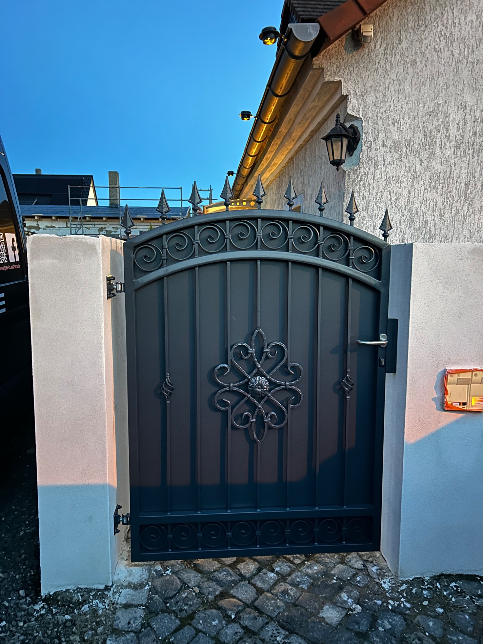 Zamknięta, ciemnoszara, metalowa furtka z ozdobnymi elementami, zwieńczona grotami, osadzona między dwoma białymi, oświetlonymi promieniami zachodzącego słońca, betonowymi słupkami, na tle...