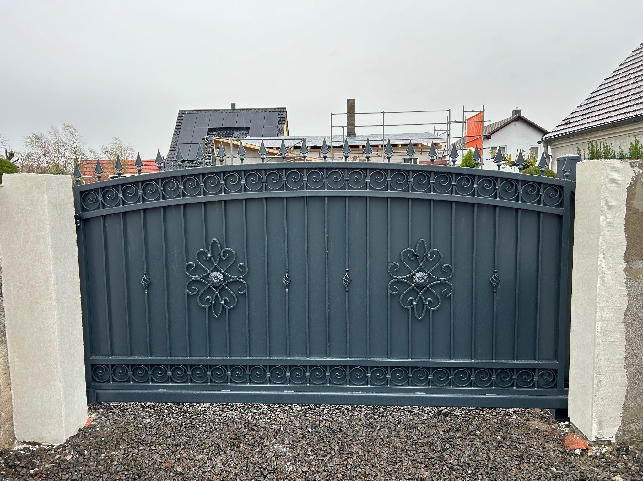 Szara brama wjazdowa z kutymi zdobieniami, osadzona między dwoma betonowymi słupkami na żwirowym podłożu, w tle zabudowa mieszkalna.