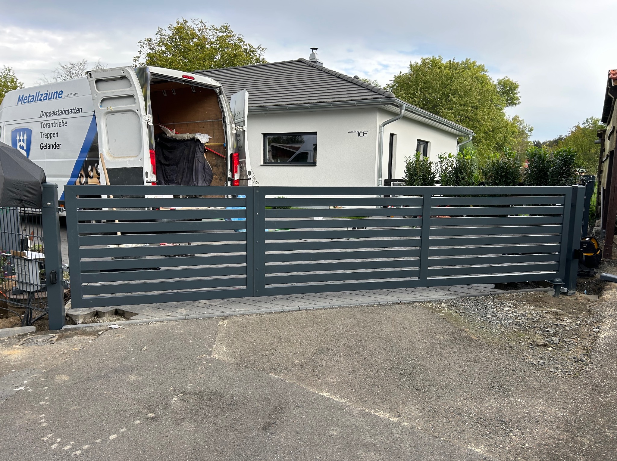 Nowoczesna, pozioma brama wjazdowa w kolorze szarym, częściowo zamontowana, z widocznym samochodem dostawczym firmy w tle oraz nowym domem jednorodzinnym.