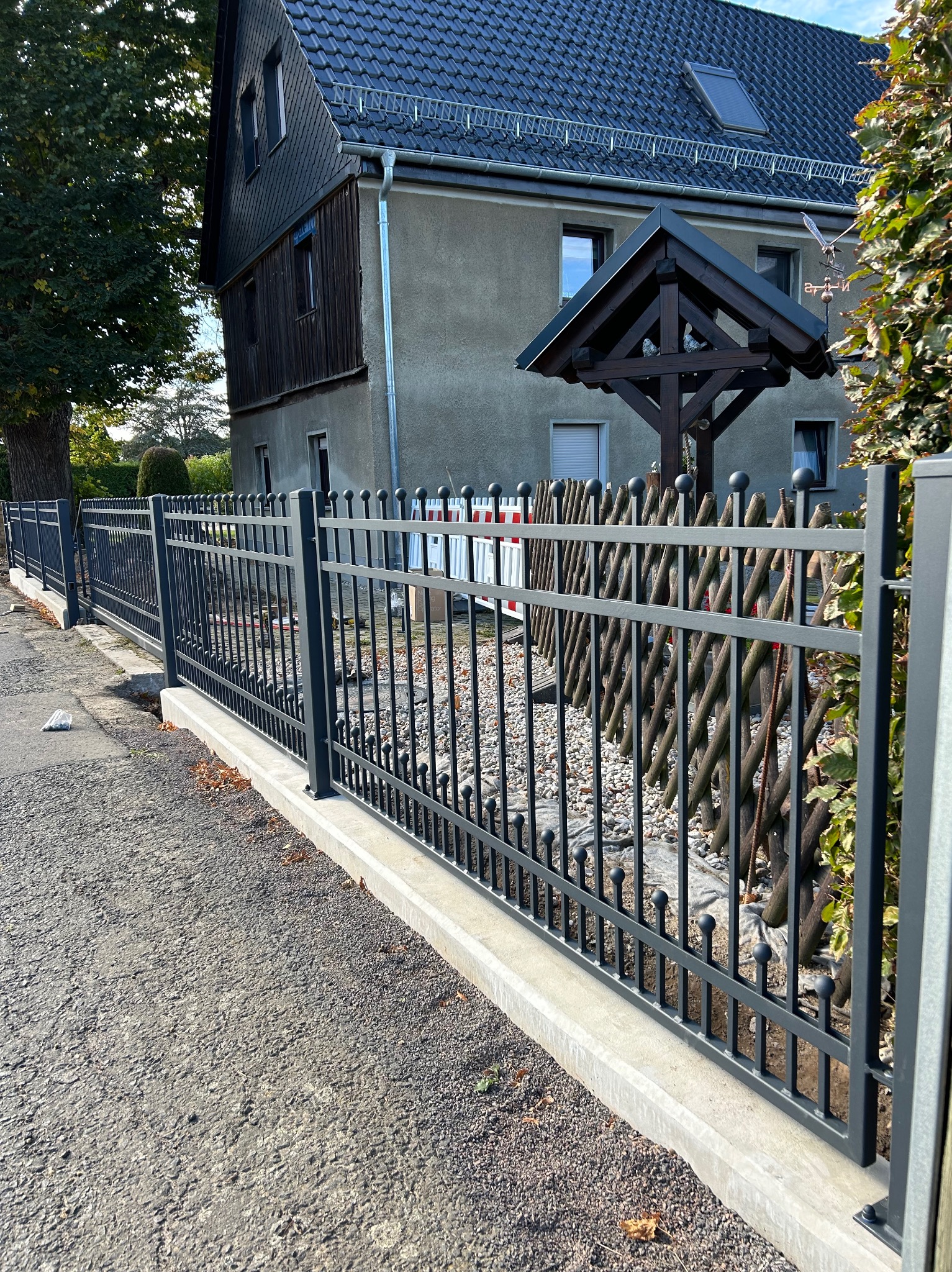 Nowoczesne, grafitowe ogrodzenie metalowe z okrągłymi zakończeniami prętów, zamontowane na betonowej podmurówce, z widocznym fragmentem posesji w tle.