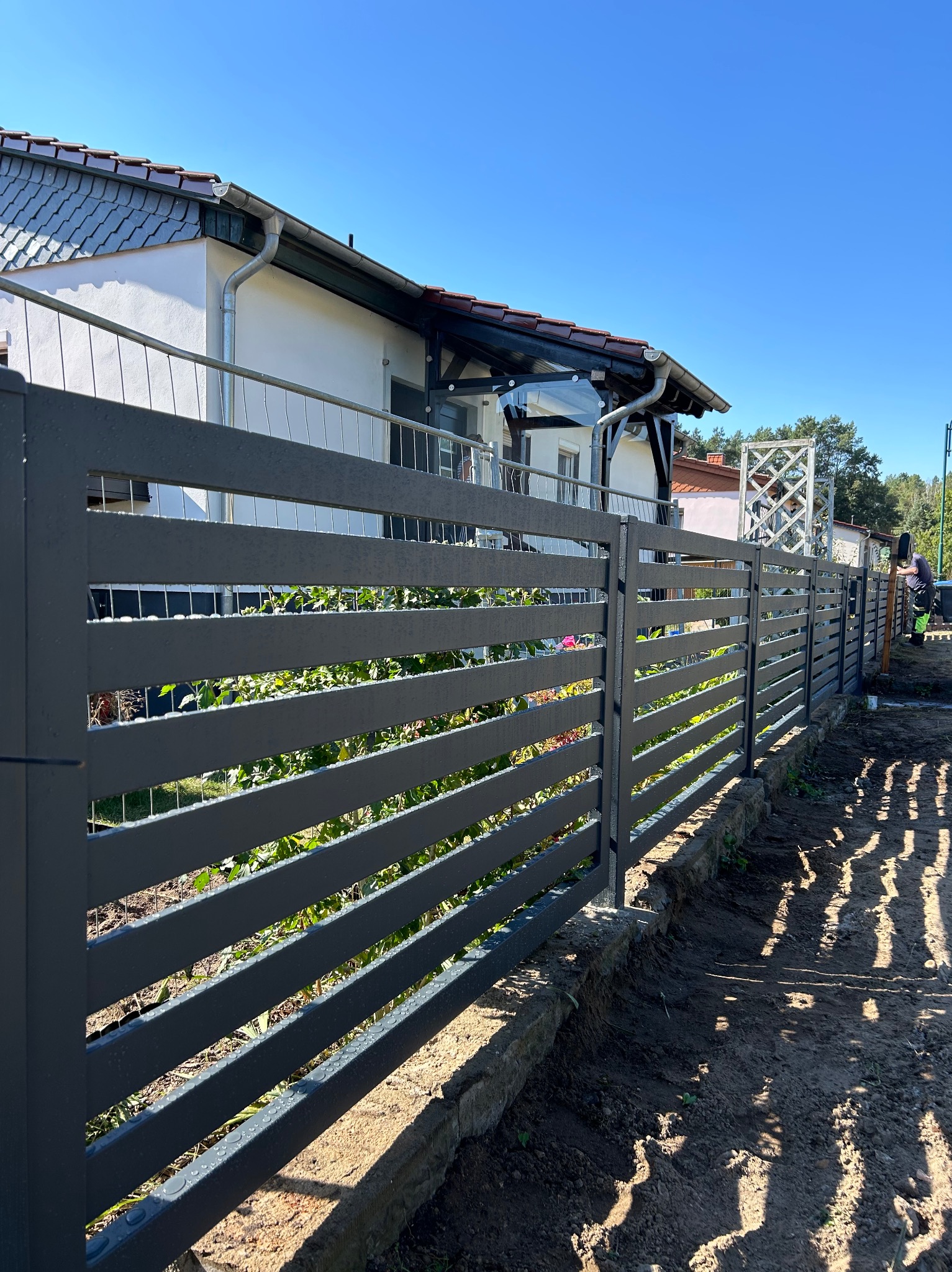 Nowoczesne, poziome ogrodzenie z szarych paneli metalowych, z widocznymi śladami po deszczu, na tle domu z białymi ścianami i zielenią ogrodu, z pracownikiem w tle.