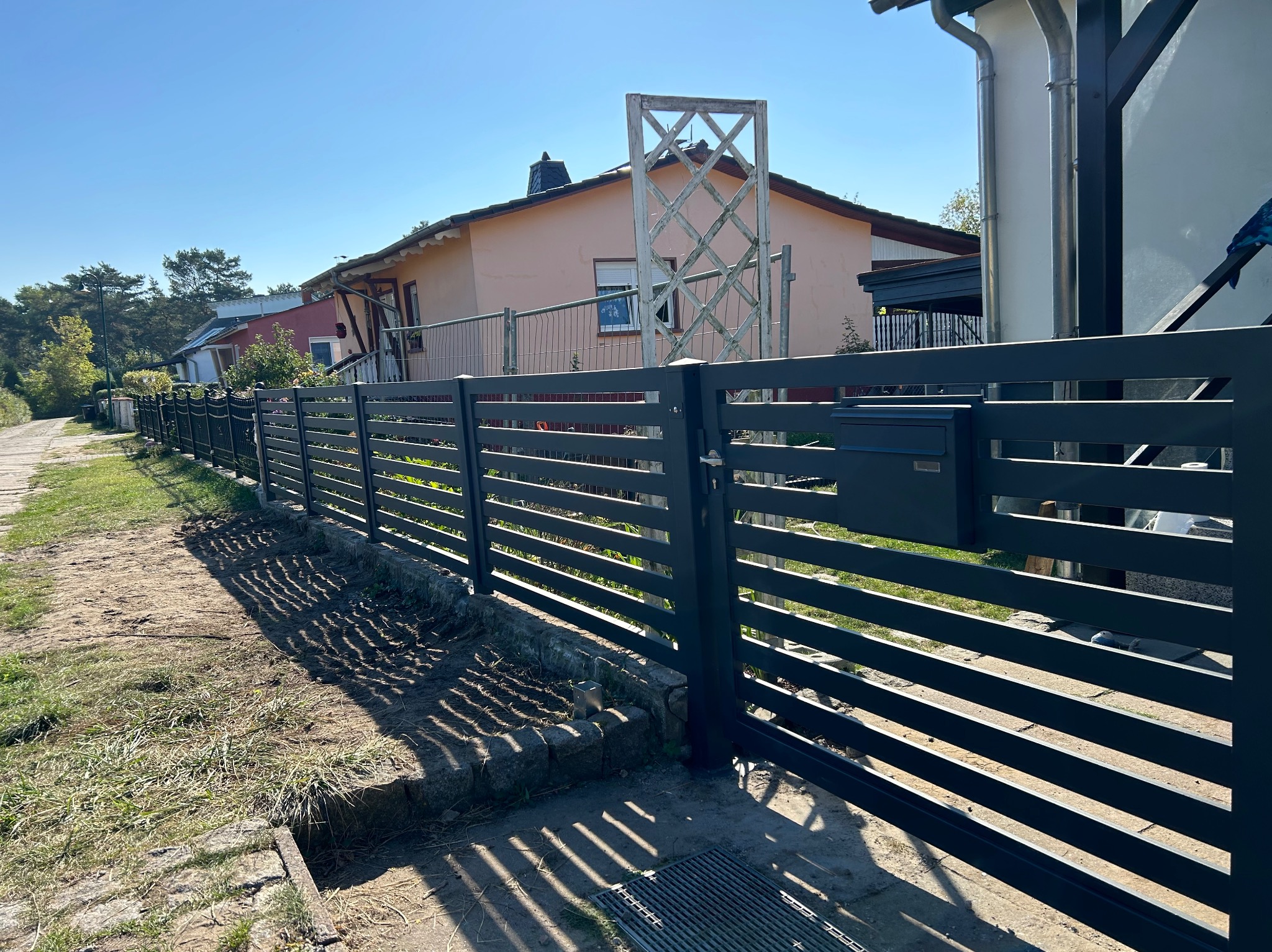 Długa, pozioma, ciemnoszara konstrukcja o regularnych prześwitach, oddzielająca posesję od chodnika, z widoczną skrzynką pocztową i otwartą furtką, w tle zabudowa jednorodzinna i błękitne niebo.