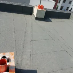 Widok z góry na płaski dach pokryty papą, z kominami wentylacyjnymi i fragmentem nowej zabudowy wielorodzinnej w tle.