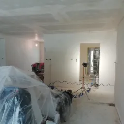 Wnętrze w trakcie prac wykończeniowych: ściany i sufit pokryte gładzią, podłoga częściowo zabezpieczona folią, widoczny sprzęt budowlany i oświetlenie przenośne.