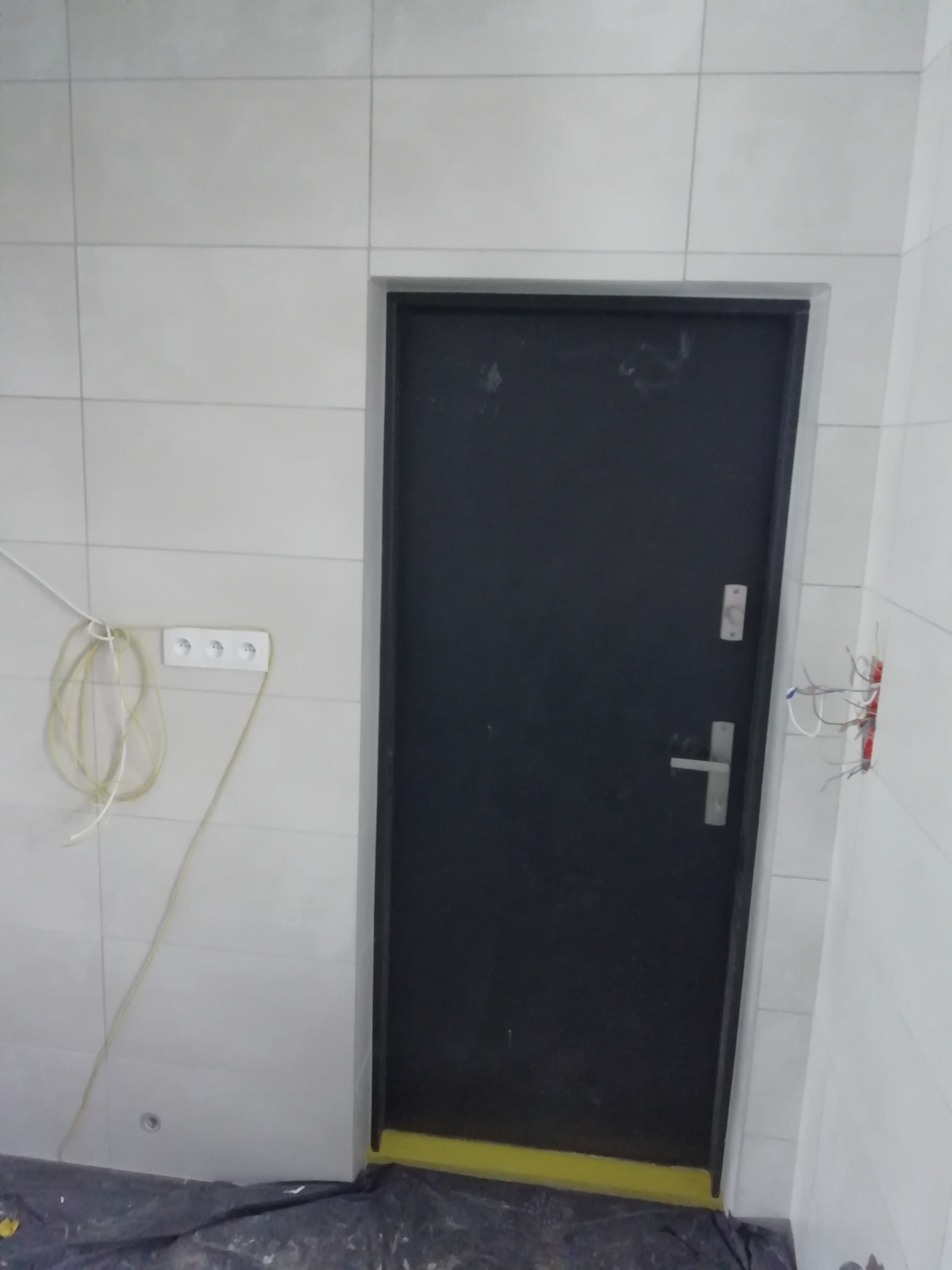 Ciemne drzwi wejściowe w trakcie montażu, widoczne białe płytki na ścianie, gniazdko elektryczne z luźno zwisającymi kablami i wystające przewody elektryczne po prawej stronie, podłoga...