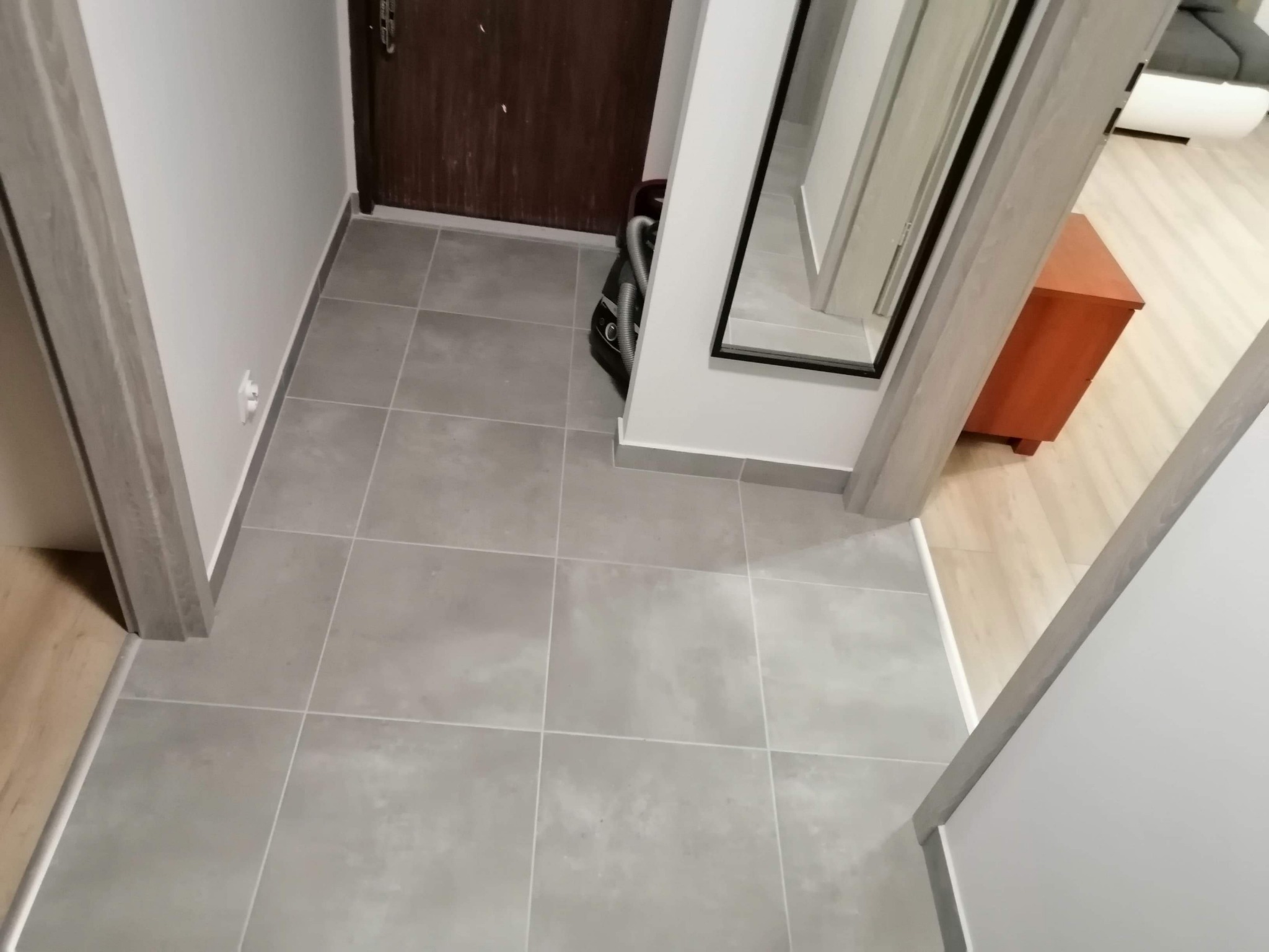 Szare, kwadratowe płytki podłogowe w przedpokoju, widoczny fragment drzwi wejściowych i lustra.