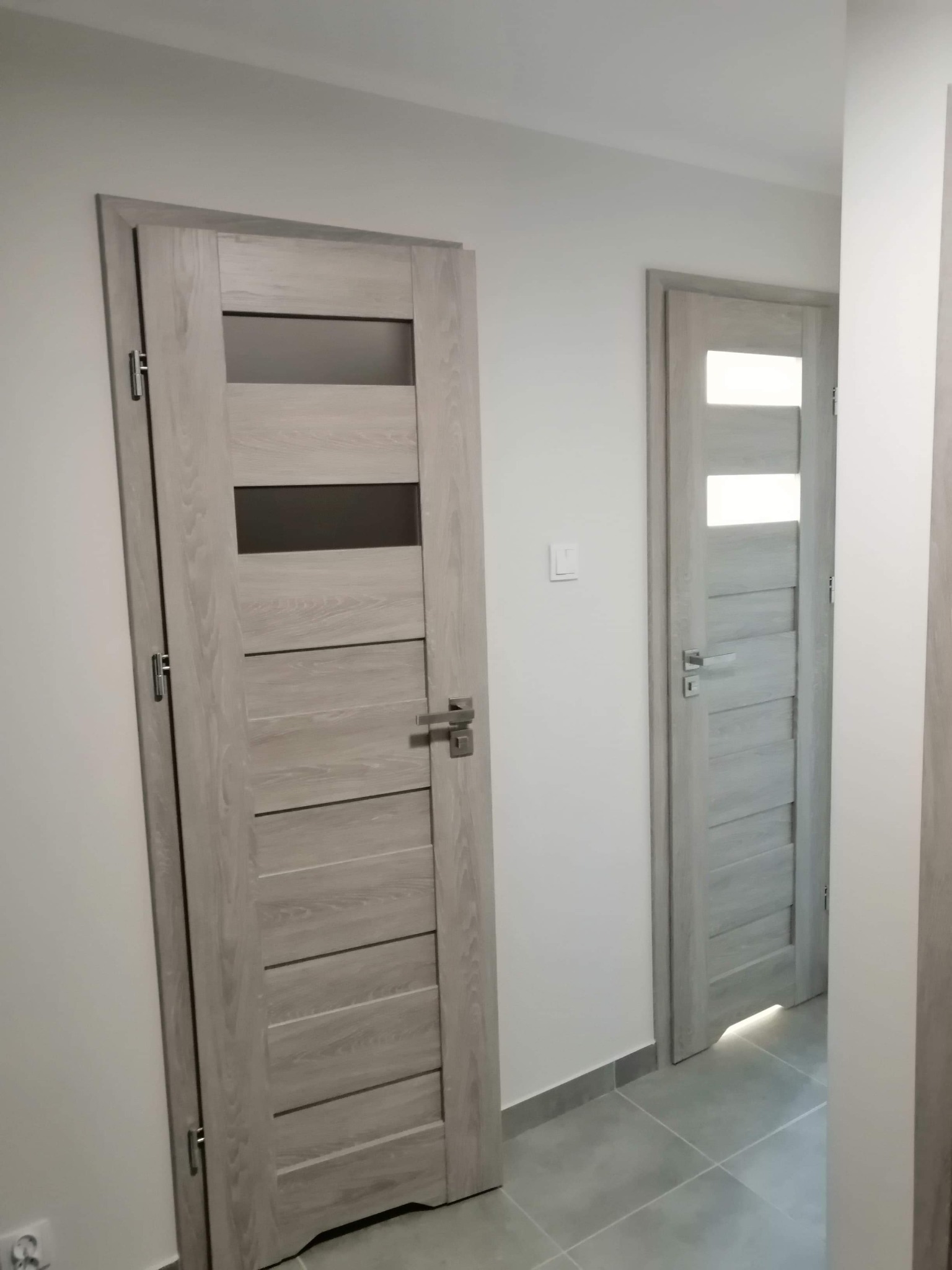 Dwa jasnoszare, drewnopodobne drzwi wewnętrzne z przeszkleniami, zamontowane w jasnym korytarzu z szarą podłogą z płytek.