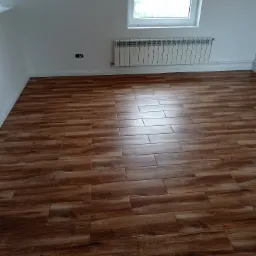 Ukończone układanie paneli podłogowych imitujących drewno w jasnym pokoju z białymi ścianami, grzejnikiem i oknem dachowym.