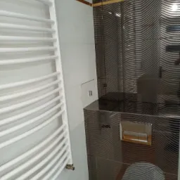 Nowoczesna łazienka z grzejnikiem drabinkowym, szare płytki z falistym wzorem i podtynkową spłuczką.