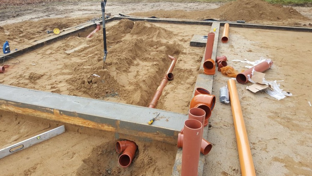 Wykopy pod fundamenty z ułożonymi rurami kanalizacyjnymi, widoczny poziomica i narzędzia, teren budowy w trakcie prac ziemnych i instalacyjnych.