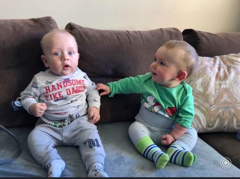 Dwaj roczni chłopcy siedzą na kanapie w Świerklańcu, jeden z nich w zielonej bluzce dotyka ramienia drugiego w szarej bluzce z napisem 'Handsome Like Daddy'.