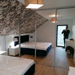 Meble Bielas - Szafa przesuwna z lustrzanymi drzwiami w sypialni na poddaszu, widoczne łóżka z folią ochronną i osoba robiąca zdjęcie w odbiciu.