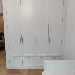 Biała szafa czterodrzwiowa z szufladami i złotymi uchwytami, widoczna w sypialni z parkietem w jodełkę i różowym żyrandolem