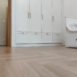 Biała szafa wnękowa z czterema drzwiami i czterema szufladami na parkiecie ułożonym w jodełkę, widok z dołu.