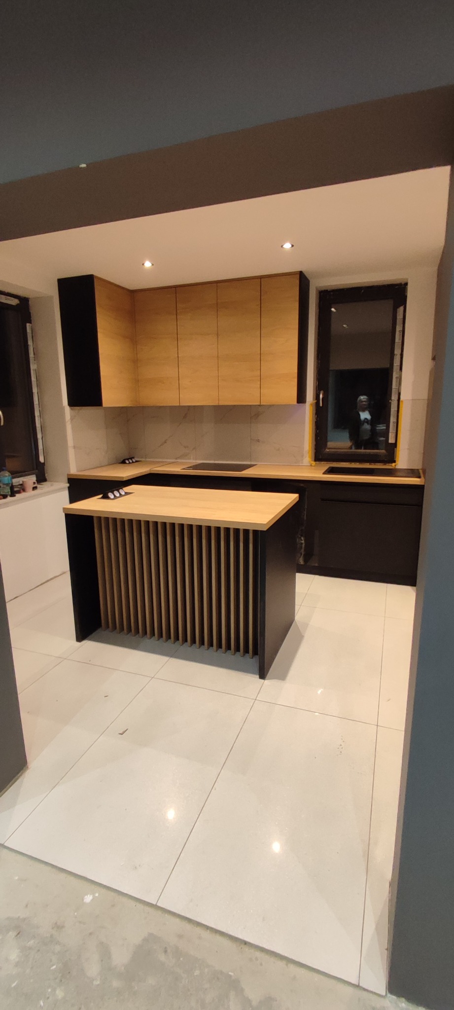 Nowoczesna kuchnia z drewnianymi frontami szafek górnych i czarnymi dolnymi, wyspa kuchenna z drewnianymi lamelami, biała marmurowa ściana nad blatem, okno z czarną ramą.