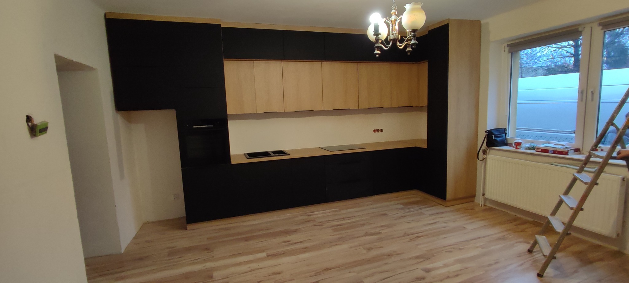 Nowoczesna kuchnia z czarnymi szafkami dolnymi i drewnianymi górnymi, widoczna drabina i akcesoria na parapecie, sugerujące montaż w toku.