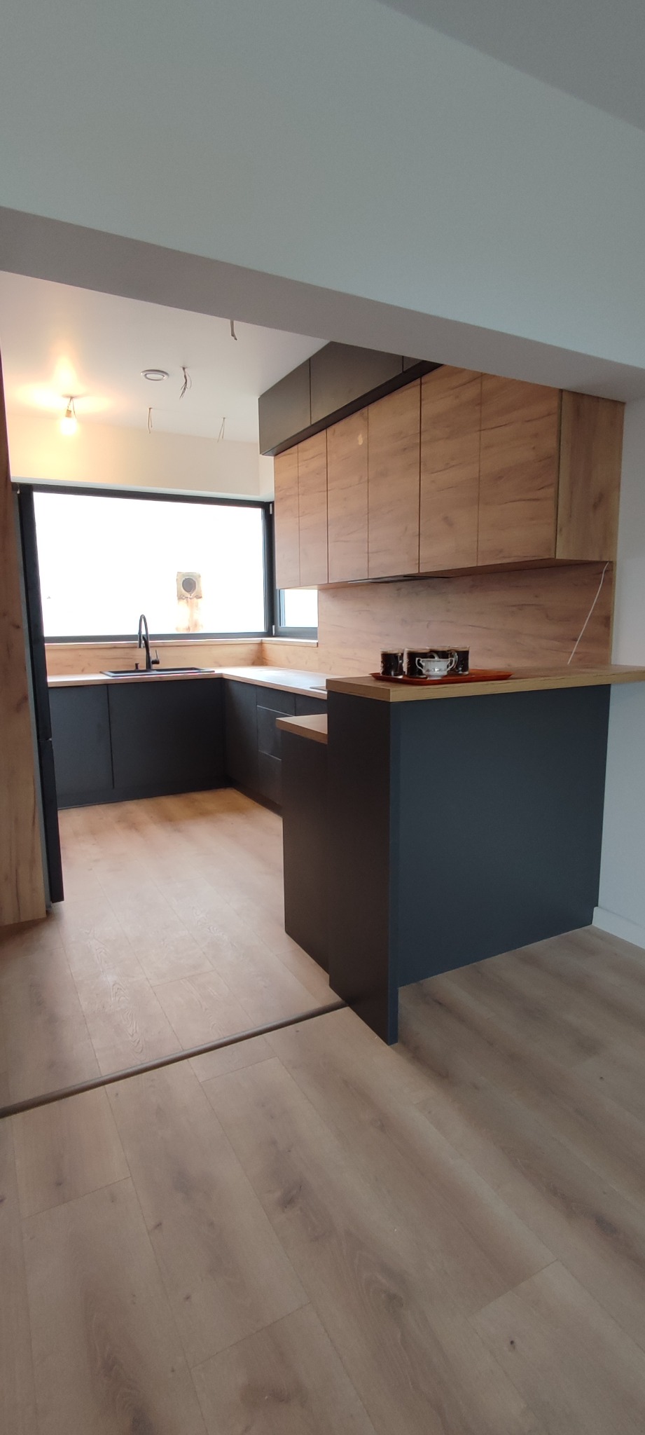 Nowoczesna kuchnia z drewnianymi frontami szafek górnych i ciemnymi szafkami dolnymi, z czarnym zlewem i baterią oraz blatem z drewna. Na blacie ekspres do kawy i filiżanki. Widoczne niedokończone...