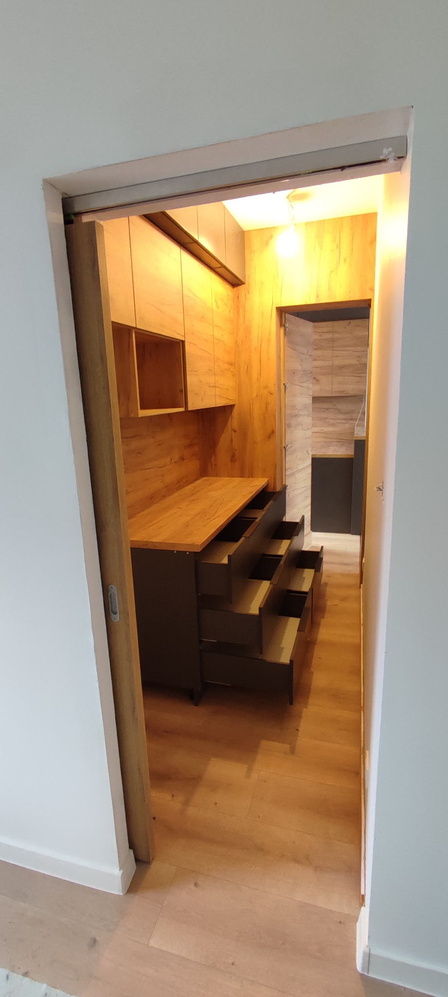 Wnętrze garderoby z drewnianą zabudową, otwarte szuflady i półki, widok z korytarza