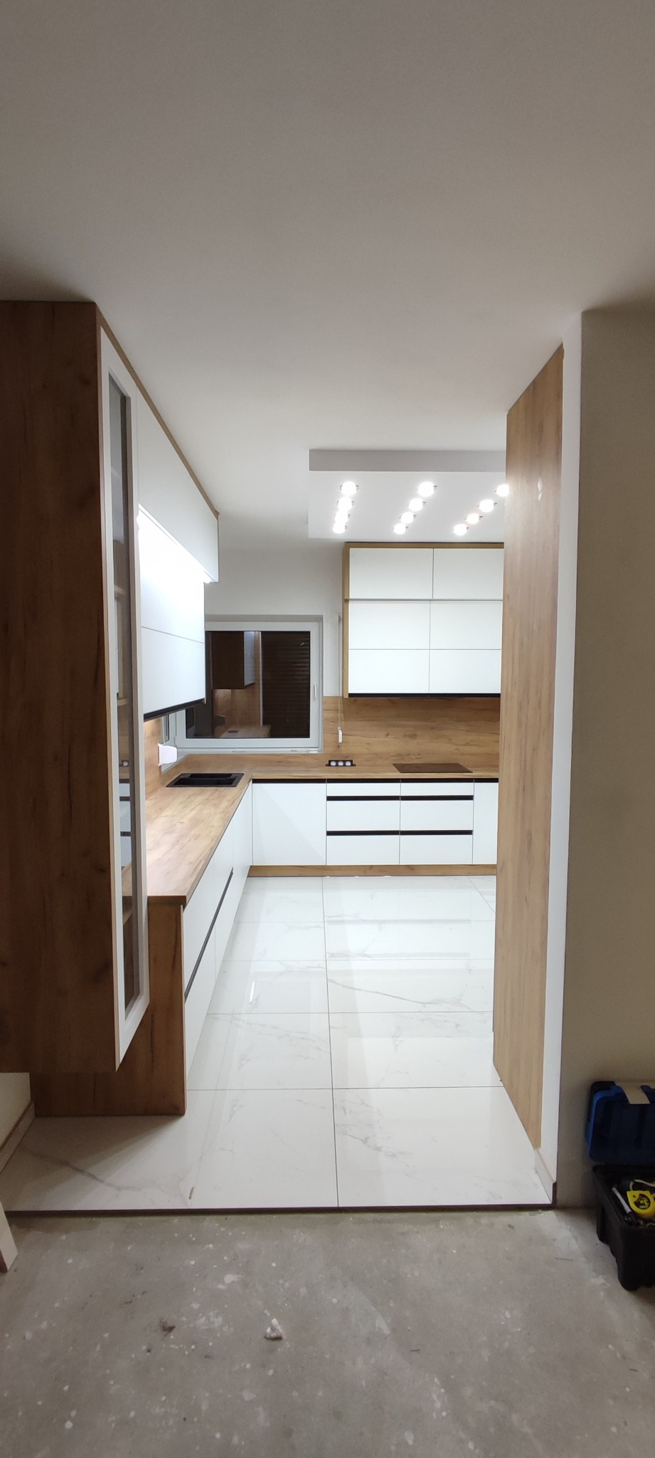 Nowoczesna kuchnia z białymi szafkami i drewnianymi blatami, widok z perspektywy wejścia, z widocznymi narzędziami na podłodze podczas montażu.