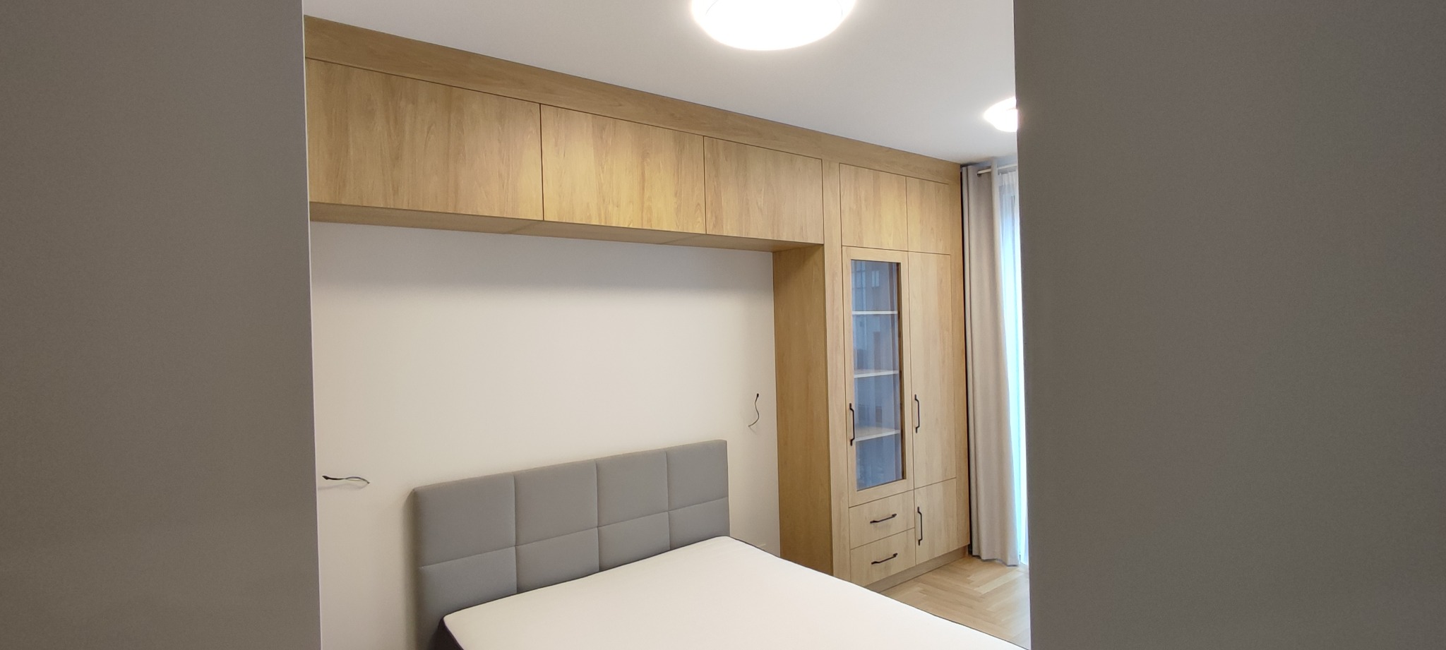 Jasna sypialnia z zabudową meblową z drewna: szafki wiszące nad łóżkiem z szarym zagłówkiem i wysoka szafa z przeszklonymi drzwiczkami.