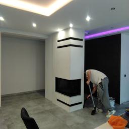 FHU Nikiel Maciej - Wnętrze po remoncie z nowoczesnym kominkiem i podświetleniem LED; pracownik sprząta podłogę po malowaniu, widoczne wiadro z farbą i narzędzia.