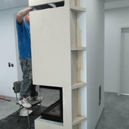 FHU Nikiel Maciej - Pracownik na krześle montuje obudowę kominka z płyty karton gips, widoczne szpachlowane łączenia i wnęki na półki.