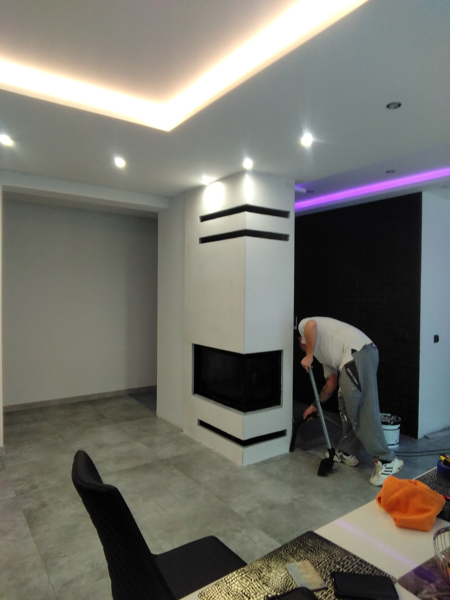 Wnętrze po remoncie z nowoczesnym kominkiem i podświetleniem LED; pracownik sprząta podłogę po malowaniu, widoczne wiadro z farbą i narzędzia.