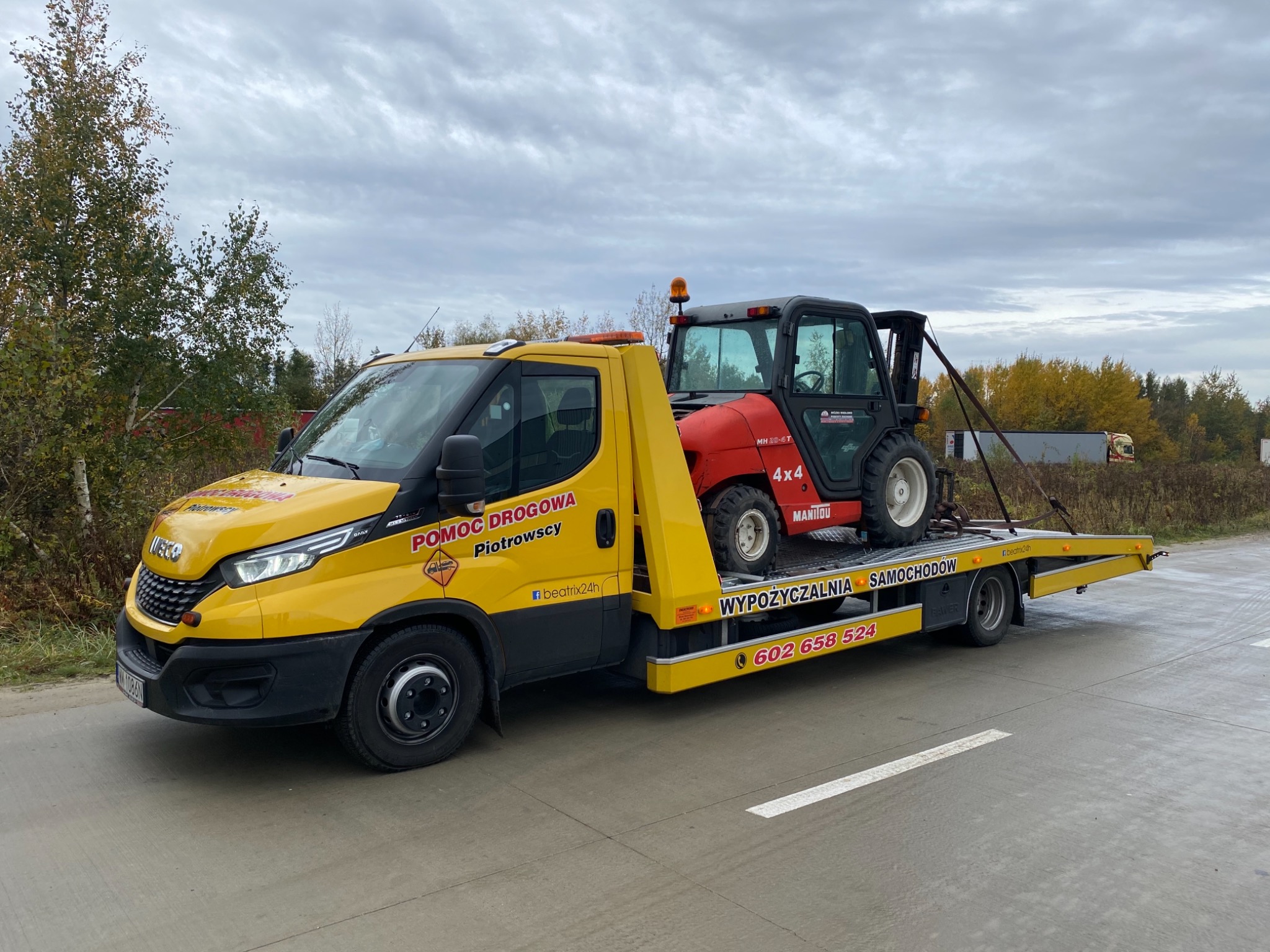 Żółta laweta pomocy drogowej z napisem 'Pomoc Drogowa Piotrowscy' przewozi czerwony wózek widłowy Manitou 4x4 na platformie, widok z boku na tle jesiennego krajobrazu.