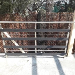 Luk-Spaw - Metalowa konstrukcja z poziomymi elementami, przypominająca barierkę lub fragment ogrodzenia, na tle siatki i bambusowej maty. Elementy poziome wykonane z szarego tworzywa, a konstrukcja nośna ze...