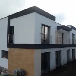 Nowoczesny dom z białymi ścianami i ciemnymi elementami wykończeniowymi, z balkonami zabezpieczonymi balustradami ze stali nierdzewnej o poziomym układzie prętów.