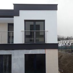 Luk-Spaw - Nowoczesny budynek z balkonem zabezpieczonym poziomymi, metalowymi barierkami na pierwszym piętrze, widok z zewnątrz na tle budynków w trakcie budowy.