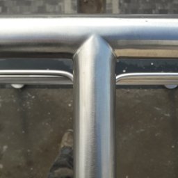 Luk-Spaw - Szczegół spawu nierdzewnej balustrady z widocznymi butami osoby fotografującej, na szarym tle chodnika.