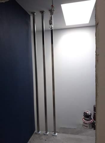 Montaż trzech pionowych, nierdzewnych słupków z kwadratowymi podstawami przy ścianie, widoczne puszki z farbą.