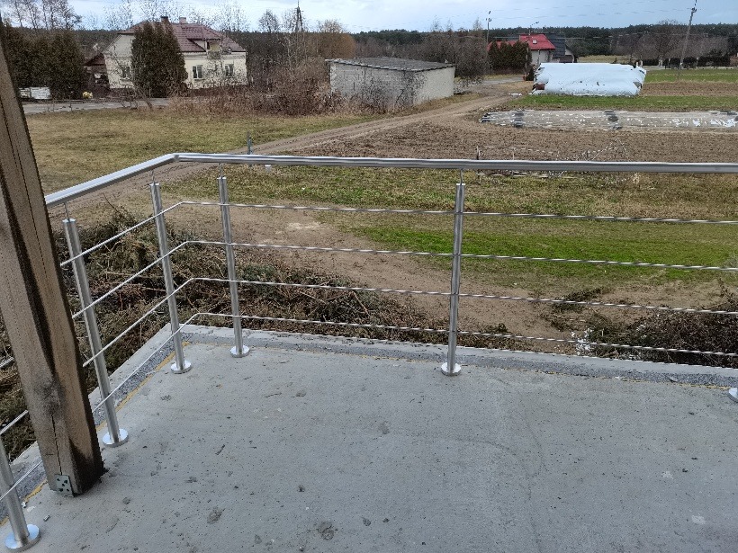 Balkon z balustradą nierdzewną o minimalistycznym designie, widok na wiejski krajobraz z domami i polami w tle.