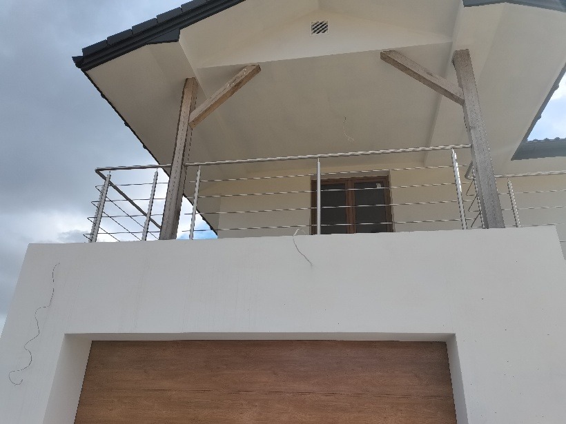 Balkon z poziomymi, metalowymi barierkami na tle białej ściany i drewnianych słupów podtrzymujących dach.