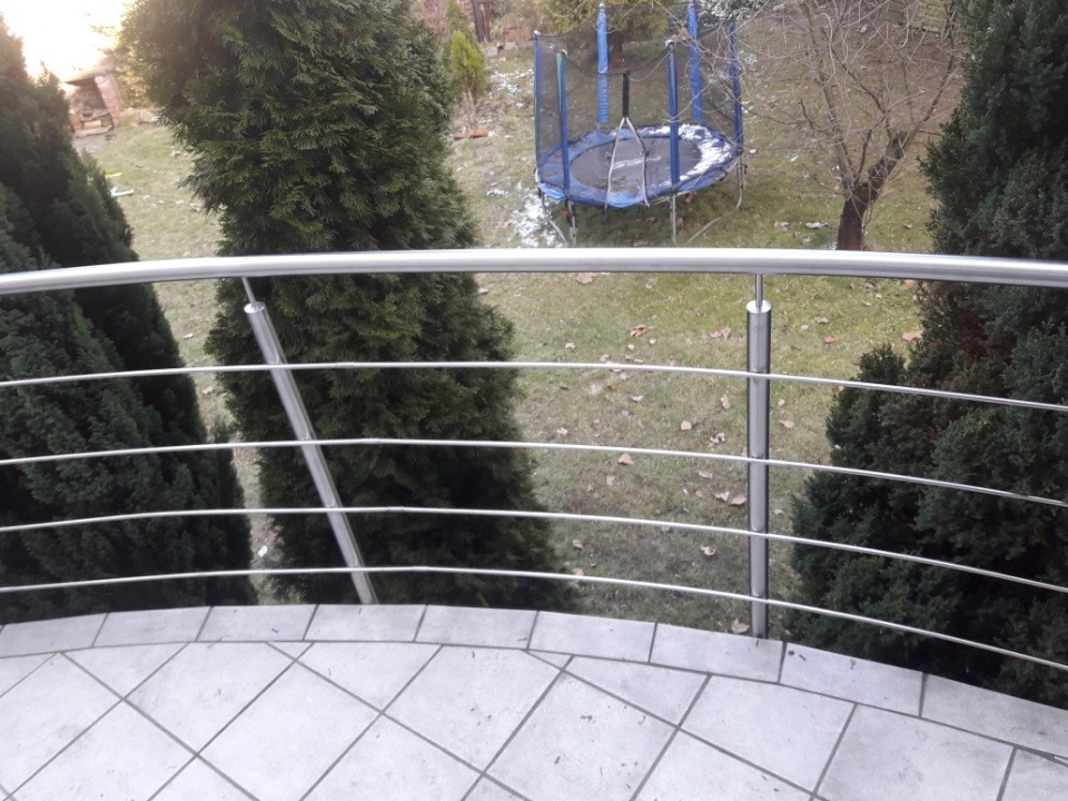 Balkon z czterema poziomami metalowych, cienkich prętów, służących jako zabezpieczenie, z widokiem na ogród z trampoliną.