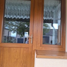 Malmar sp. z .o.o - Brązowe, drewniane drzwi balkonowe z szybami, przez które widać firankę i zieleń. Zewnętrzny parapet w kolorze drzwi. Widoczny fragment elewacji.