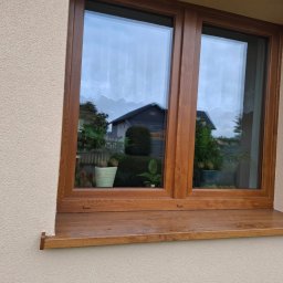 Malmar sp. z .o.o - Dwuskrzydłowe okno PCV w kolorze drewna, z widocznymi roślinami w doniczkach na parapecie i odbiciem domu w szybie, osadzone w elewacji o fakturze baranka.