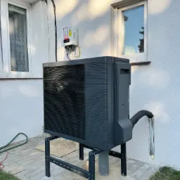 Czarna pompa ciepła na metalowym stelażu, zamontowana przy białej ścianie domu z dwoma oknami, widoczny falownik fotowoltaiczny i przewody.