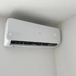 Biała klimatyzacja marki AUX zamontowana na jasnej ścianie, wyświetlacz pokazuje temperaturę 20 stopni Celsjusza, widoczny fragment futryny drzwi po lewej stronie.