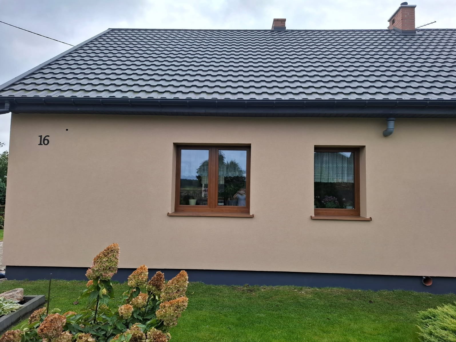 Dom jednorodzinny z beżową elewacją, ciemną dachówką i numerem 16. Widoczne dwa okna z drewnianą ramą i fragment ogrodu z hortensjami.