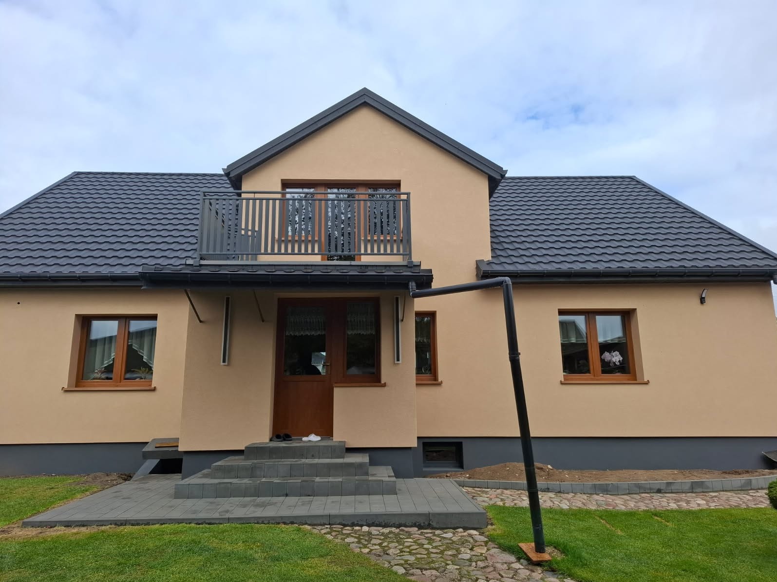 Dom jednorodzinny z beżową elewacją, grafitowym dachem i balkonem z metalową balustradą. Widoczne okna z drewnianą ramą i schody wejściowe z szarej kostki.