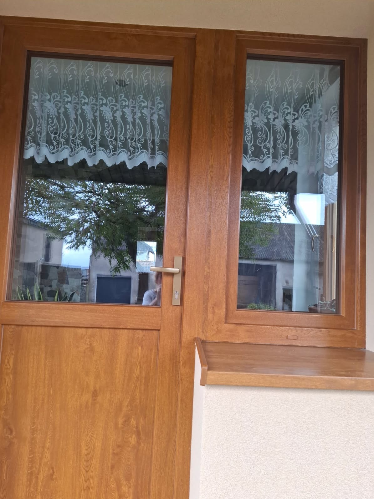 Brązowe, drewniane drzwi balkonowe z szybami, przez które widać firankę i zieleń. Zewnętrzny parapet w kolorze drzwi. Widoczny fragment elewacji.