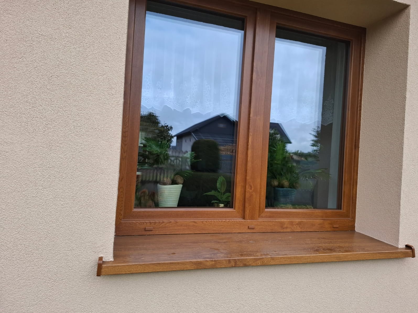 Dwuskrzydłowe okno PCV w kolorze drewna, z widocznymi roślinami w doniczkach na parapecie i odbiciem domu w szybie, osadzone w elewacji o fakturze baranka.