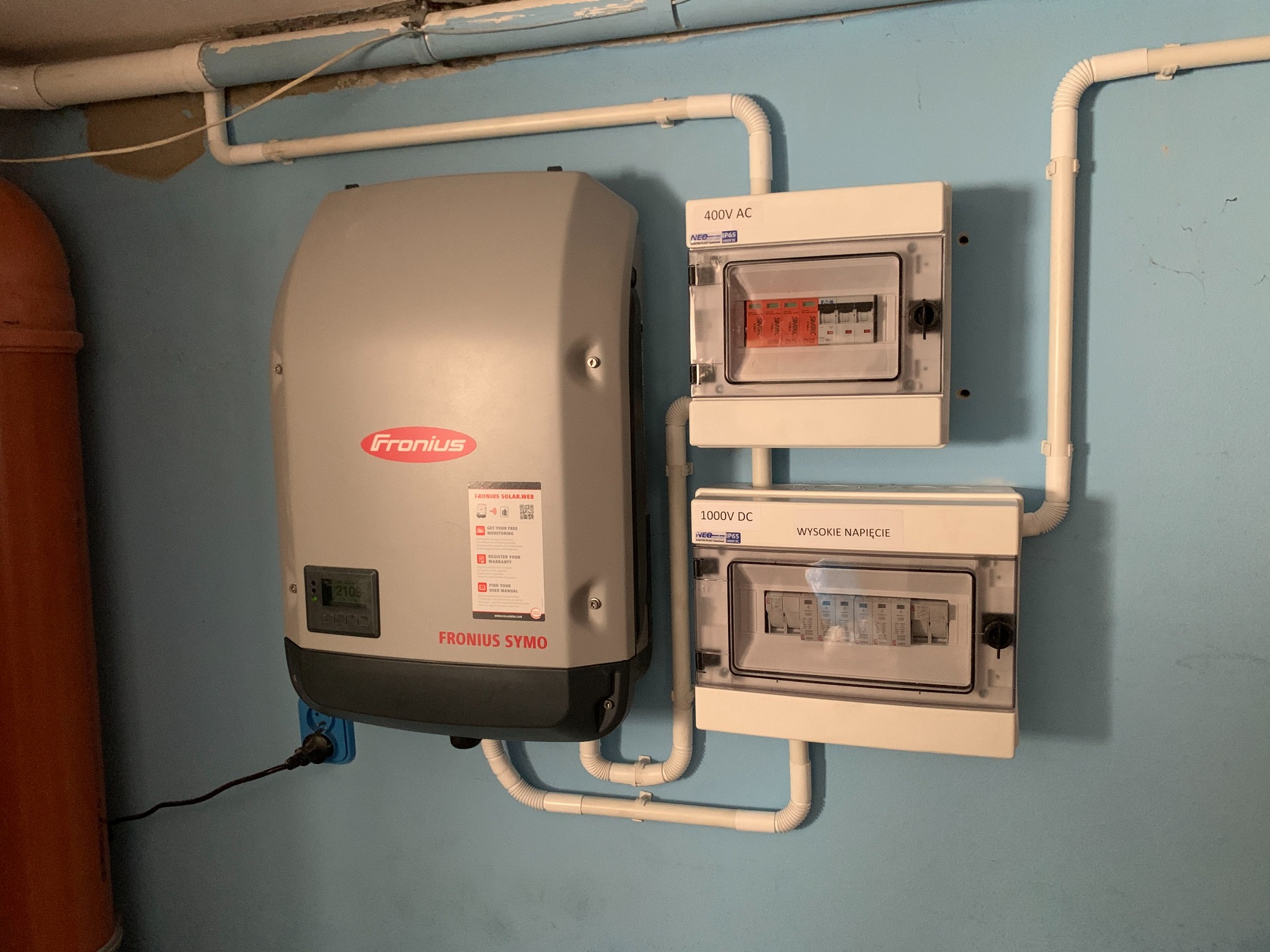Szary falownik Fronius Symo zamontowany na niebieskiej ścianie obok dwóch białych skrzynek z bezpiecznikami, połączony białymi rurkami PCV. Widoczne oznaczenia 400V AC i 1000V DC.