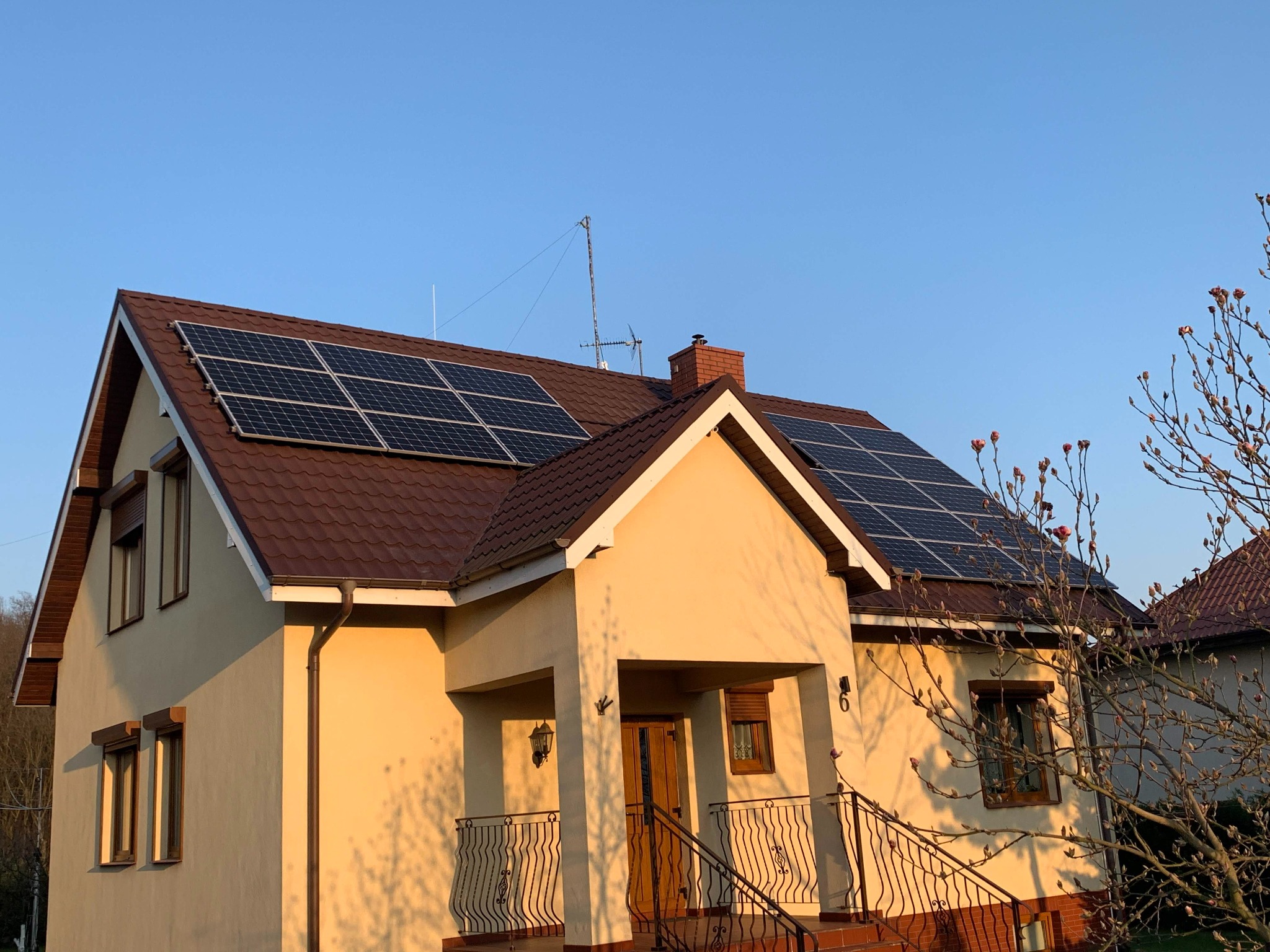 Dom jednorodzinny z brązowym dachem, na którym zamontowane są panele słoneczne; widoczne drzewo z pąkami kwiatowymi i błękitne niebo w tle.