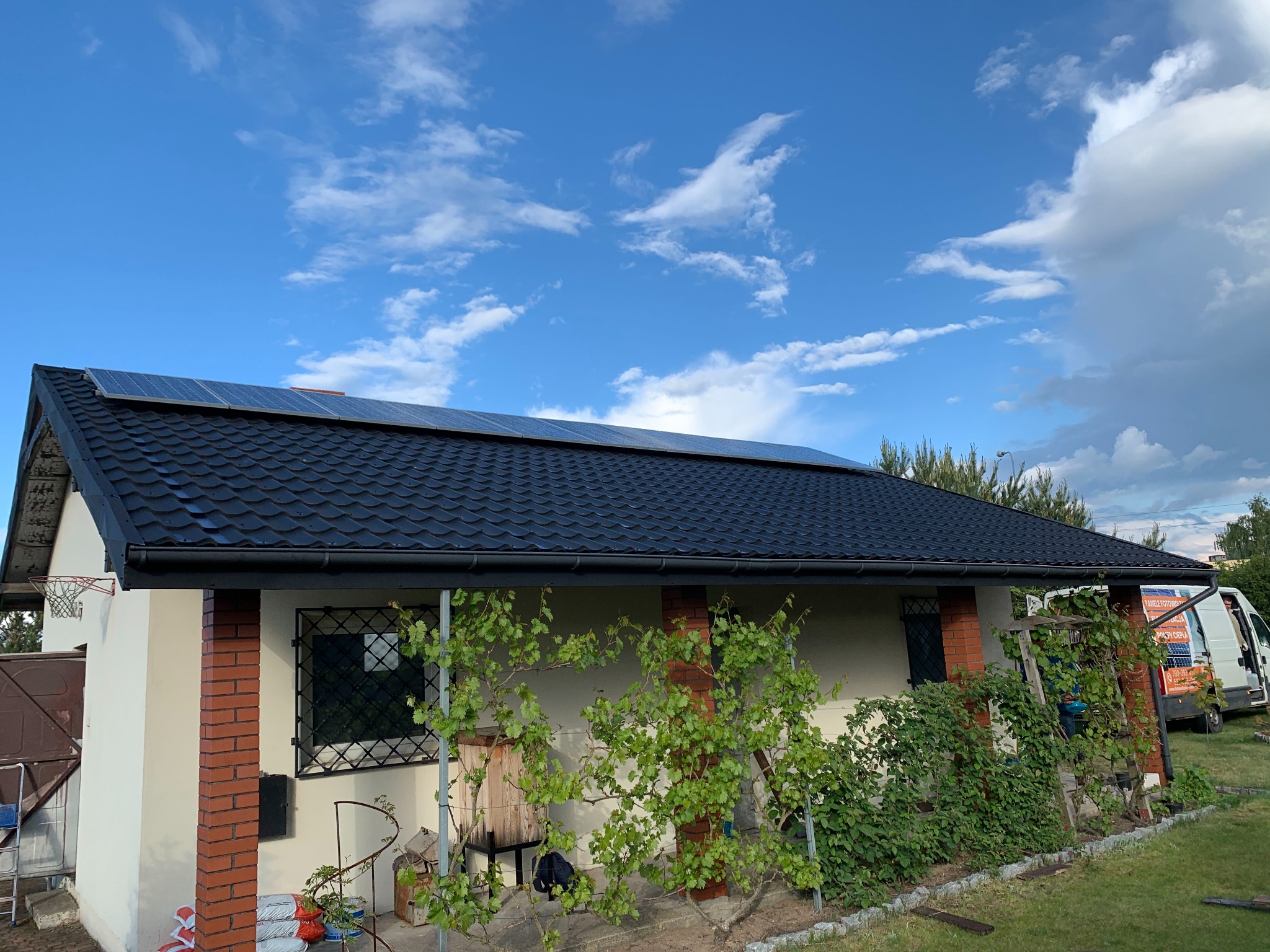 Dom jednorodzinny z panelami słonecznymi na dachu pokrytym czarną dachówką, częściowo zasłonięty pnączami, na tle błękitnego nieba z obłokami.