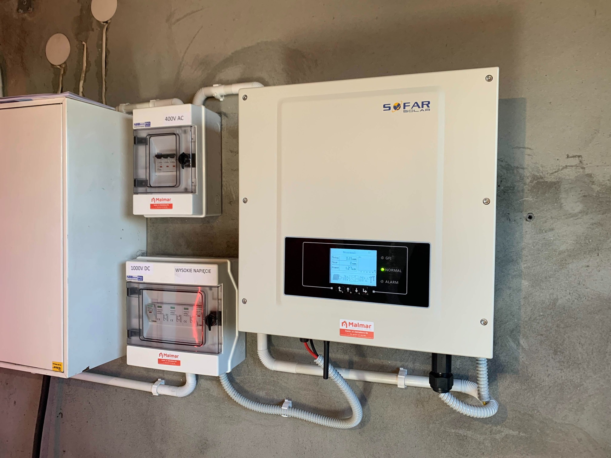 Biały inwerter SOFAR Solar zamontowany na surowej, betonowej ścianie, obok skrzynki z bezpiecznikami 400V AC i 1000V DC, instalacja elektryczna z rurkami ochronnymi.