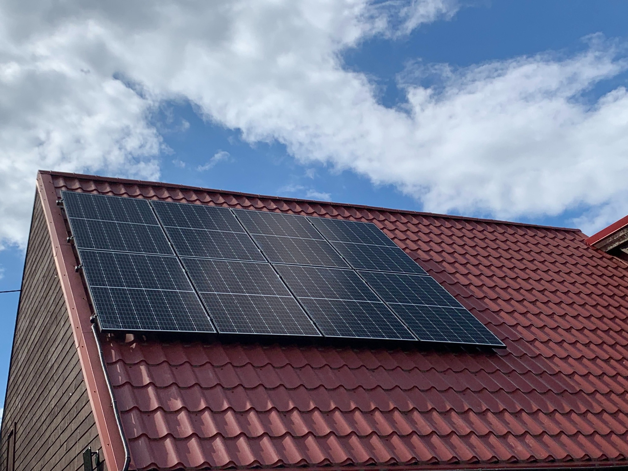 Panele słoneczne zamontowane na dachu z czerwonej dachówki, widok z dołu na tle błękitnego nieba z chmurami.